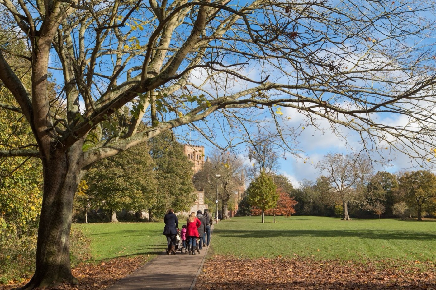 Walkers on a tree-lined park path below the cathedral tower on an autumn afternoon
